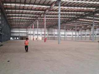 Large empty warehouse interior with concrete floor, steel columns, roof trusses, roller shutters, fire equipment and a worker in a safety vest.
