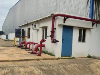 Exterior of a corrugated warehouse with a small white utility room, blue door, red fire pipes, water tank and paved yard.