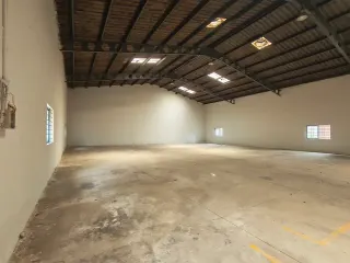Empty warehouse interior with concrete floor, white walls, small windows, electrical wiring, and corrugated metal roof trusses.