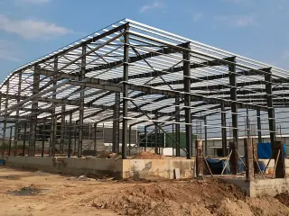 Exterior view of a steel-frame warehouse under construction with partial roof sheets, concrete foundation, dirt ground and blue sky.