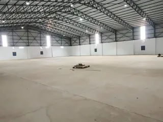 Empty warehouse interior with a concrete floor, white walls, steel roof trusses, skylight panels, and a few small objects on the floor.