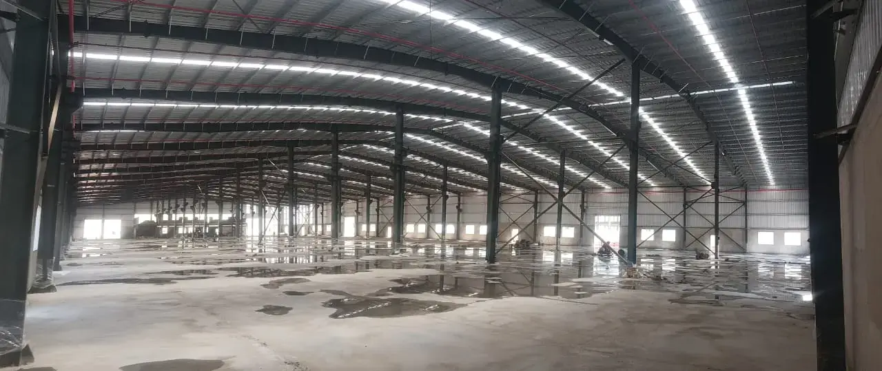 Large empty warehouse interior with steel columns, roof trusses, strip lights, and a concrete floor with puddles.