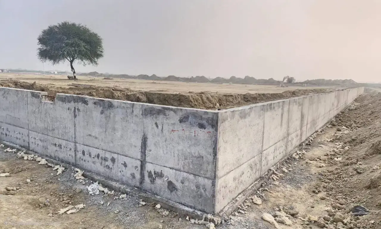 Concrete boundary or foundation wall surrounding an open dirt plot, with a lone tree and earthworks in the background.