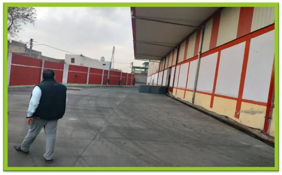 Paved yard beside a long warehouse exterior with red-and-white wall panels and canopy; a man walks toward a red boundary wall.