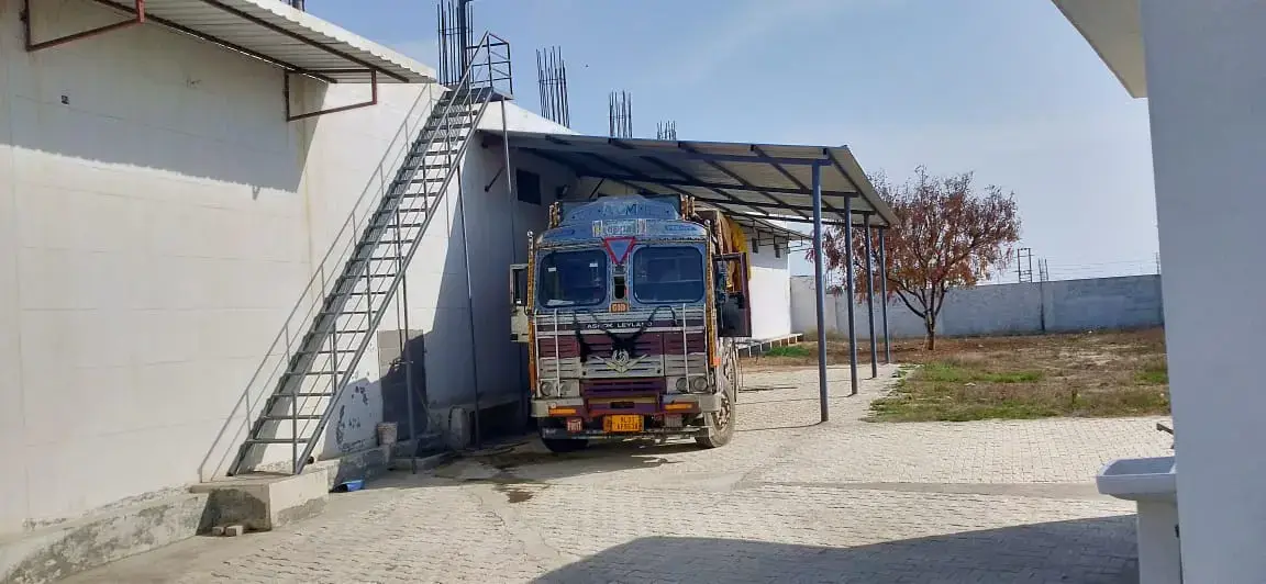Truck parked beside a warehouse wall with an exterior metal staircase, canopy, paved yard and open land beyond.